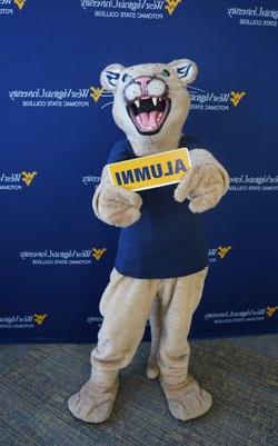 a picture of the wvu potomac state college mascot posing for a picture, holding a sign with the word "ALUMNI" on it